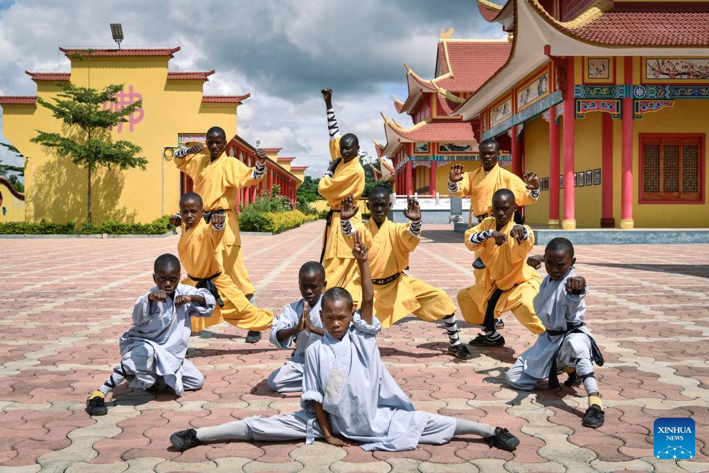 A group of young Shaolin students at the Zambia Cultural Centre pose in martial arts stances in the courtyard, some in yellow training uniforms and some in white, with the temple's red and gold pagoda buildings behind them.