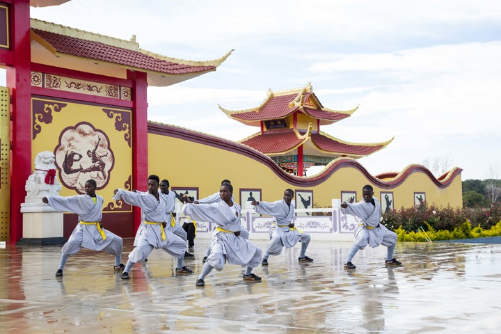 Shaolin disciples in white training uniforms perform a staff-form drill in unison in the courtyard of the Shaolin Temple Cultural Centre, Lusaka, with the red and gold entrance gate behind them.